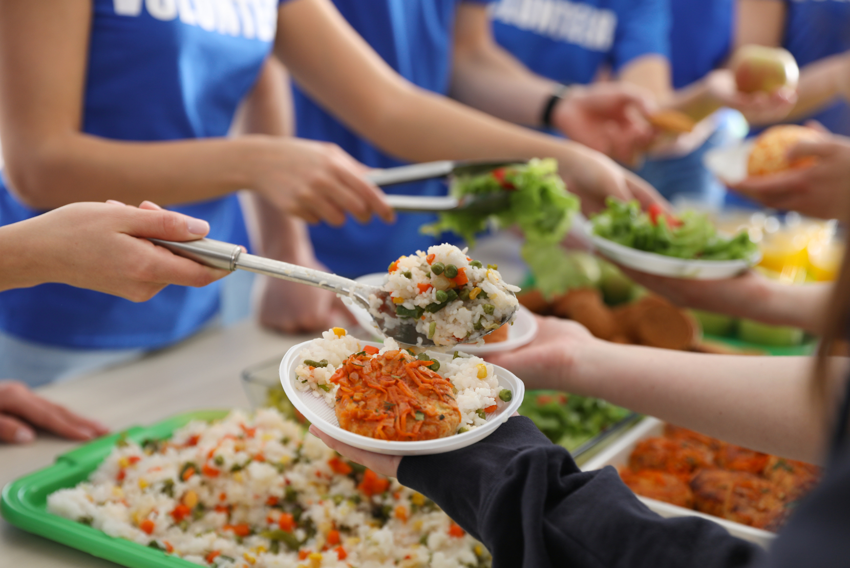 People in blue shirts serve themselves at a buffet, scooping mixed rice with vegetables onto plates.